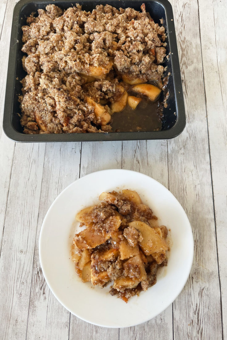 Dairy-free apple crumble on a white plate with a pan of the apple crumble in the background, all on a white wood table.