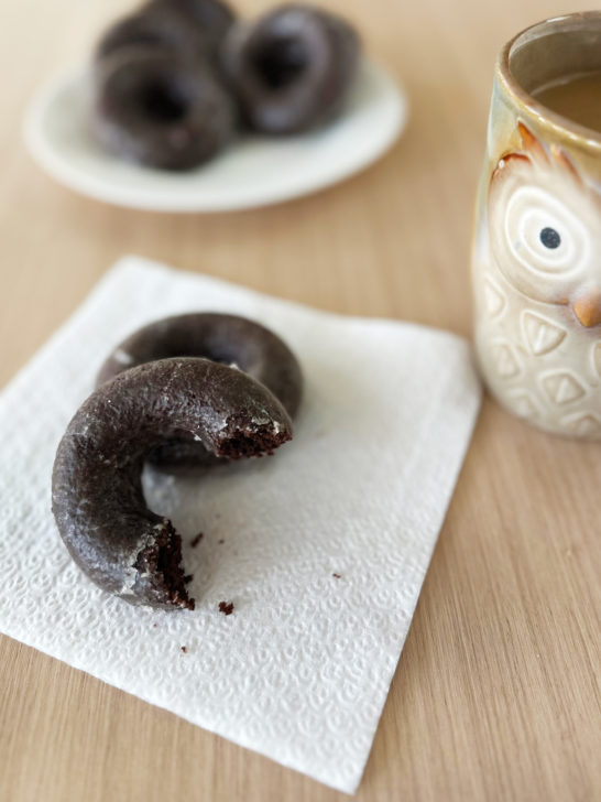 Baked Chocolate Donuts on a napkin with more on a plate near a cup of coffee on a light wood table.