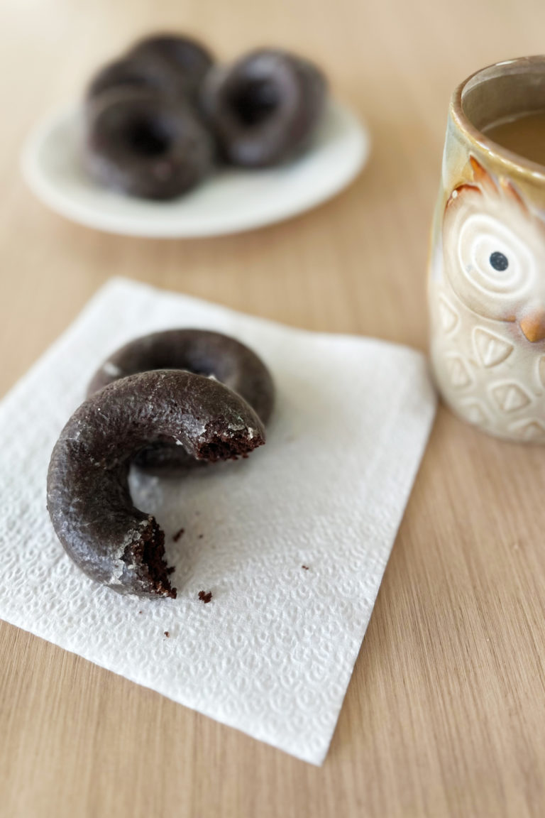 Dairy-free chocolate doughnuts with a shiny glaze piled on a white plate and in front is a napkin with a donut and a half on a napkin next to a mug of coffee.