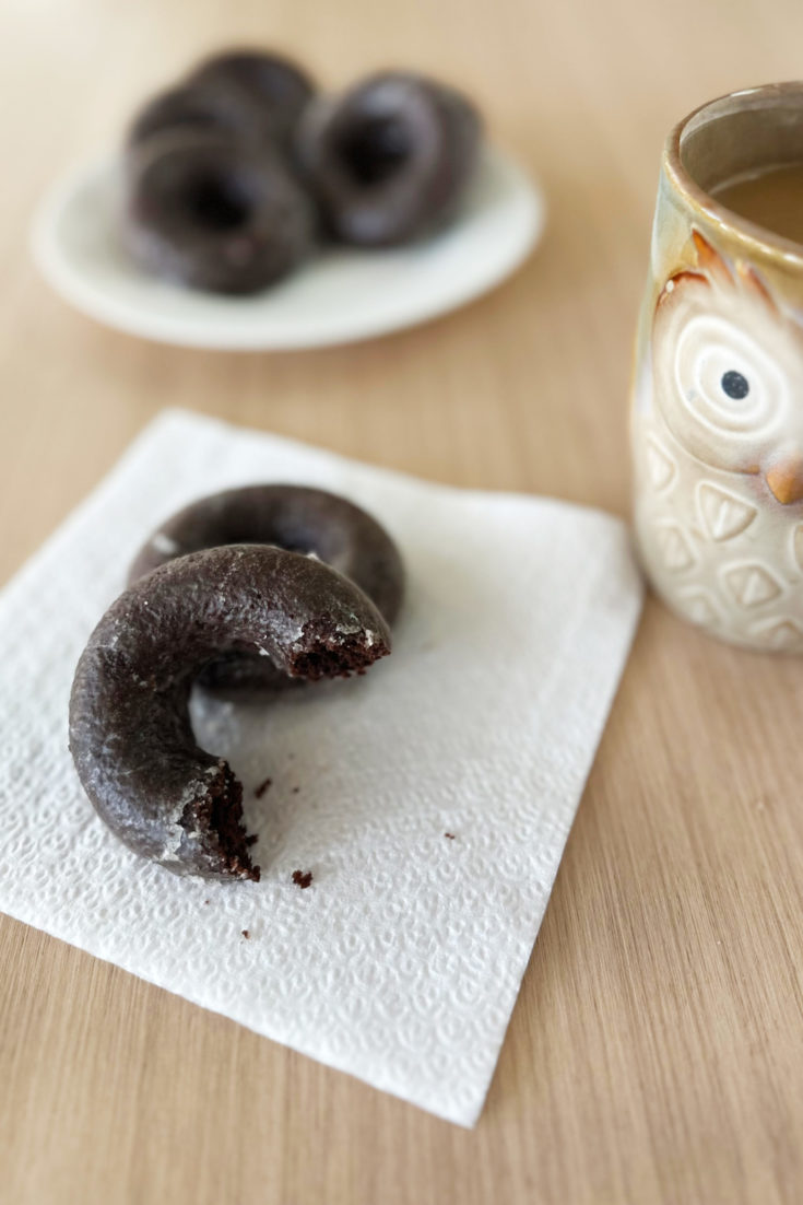 Baked Chocolate Donuts on a napkin with more on a plate near a cup of coffee on a light wood table.