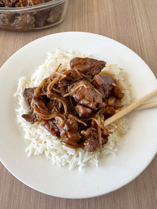 Kosher chop suey over rice with chopsticks on a white plate on a light wood background.