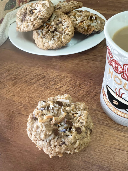 dairy-free cowboy cookies on a white plate with a towel nearby and a cup of coffee next to a cookie on a dark wood table.