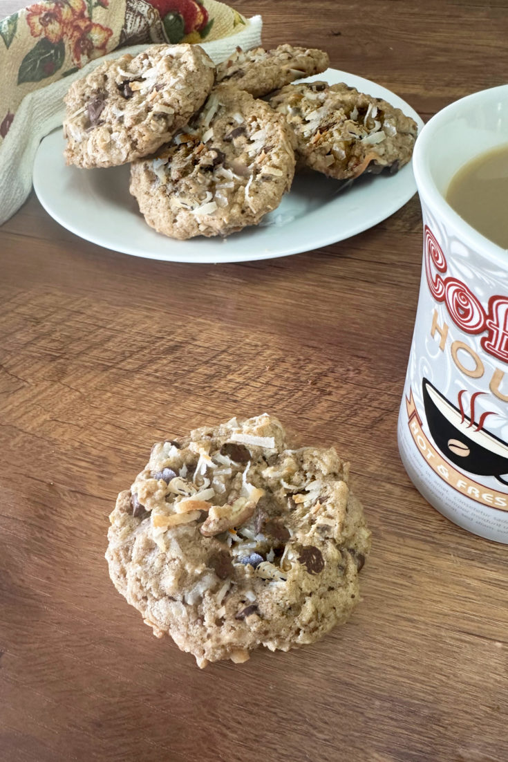dairy-free cowboy cookies on a white plate with a towel nearby and a cup of coffee next to a cookie on a dark wood table.