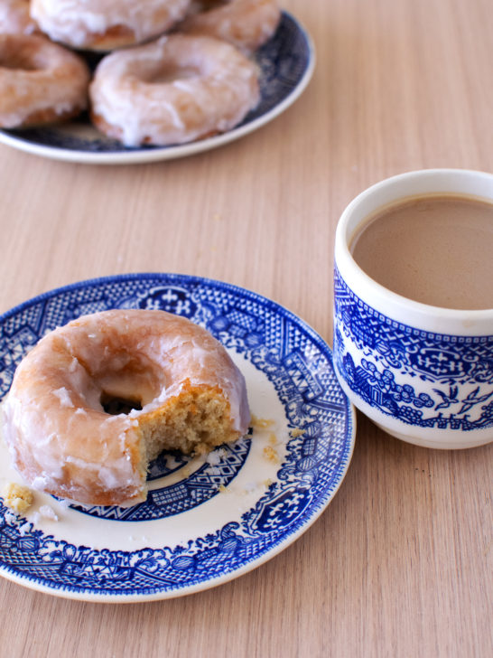 dairy-free baked cake donuts on a blue plate in the background and one donut on a blue plate in front with a bite taken out and a cup of coffee nearby, all on a wood table.