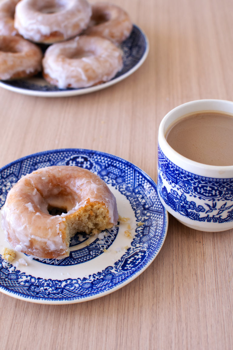 dairy-free baked cake donuts on a blue plate in the background and one donut on a blue plate in front with a bite taken out and a cup of coffee nearby, all on a wood table.