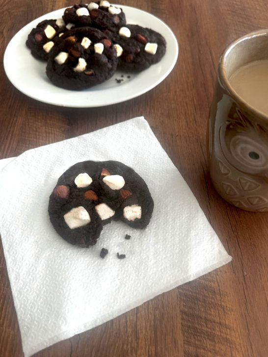 Rocky road cookies on a white plate with a cookie missing a bite on a napkin in front of it and a cup of coffee to the right, all on a dark wood tabl.e