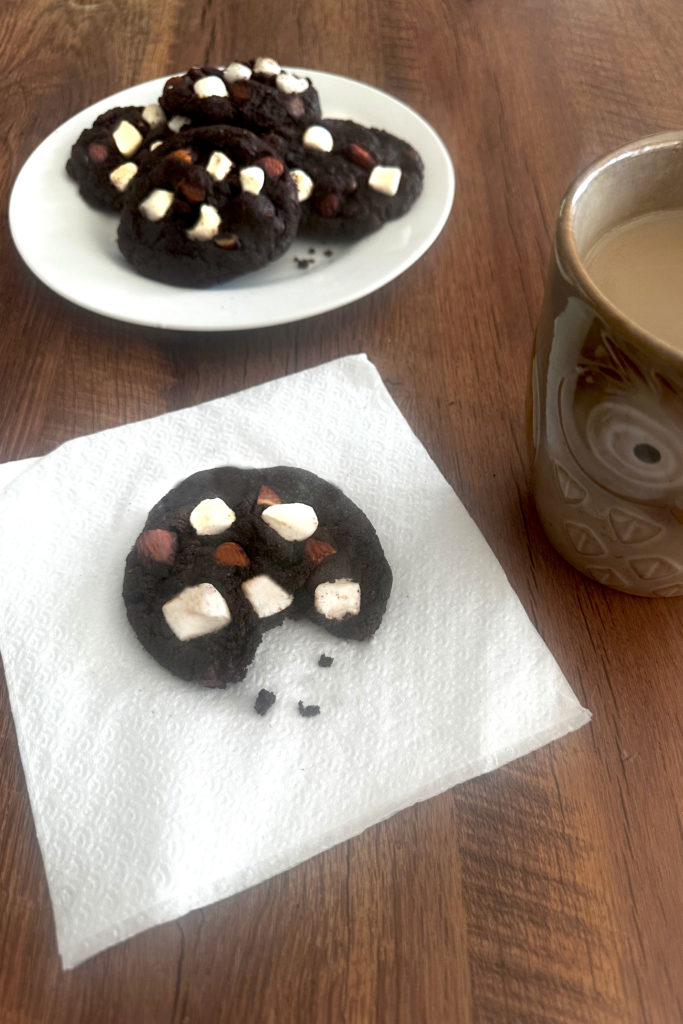 Rocky road cookies on a white plate with a cookie missing a bite on a napkin in front of it and a cup of coffee to the right, all on a dark wood tabl.e