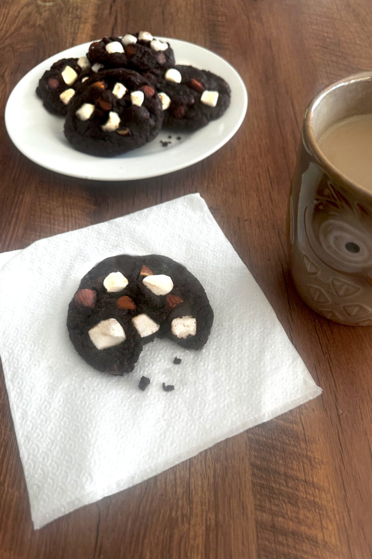 Rocky road cookies on a white plate with a cookie missing a bite on a napkin in front of it and a cup of coffee to the right, all on a dark wood tabl.e