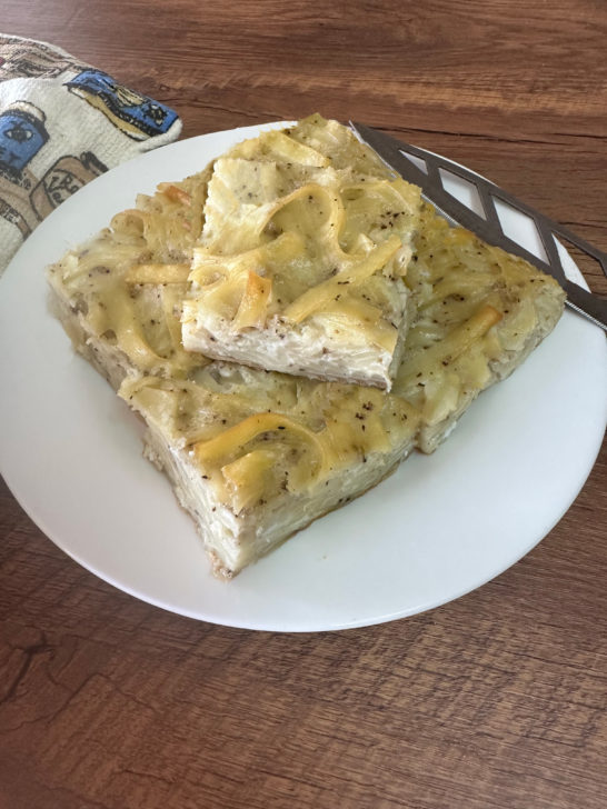 Pieces of Salt and pepper kugel on a white plate with a knife and a towel nearby on a dark wood table