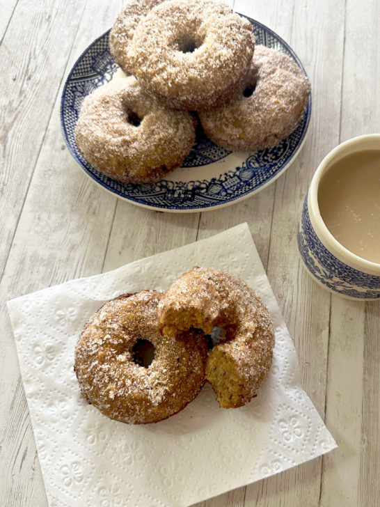 Baked apple doughnuts on a blue plate in the background an in front is a napkin with a donut and a half on it with a cup of coffee nearby, all on a white wood table
