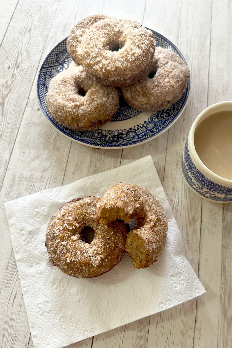Baked apple doughnuts on a blue plate in the background an in front is a napkin with a donut and a half on it with a cup of coffee nearby, all on a white wood table