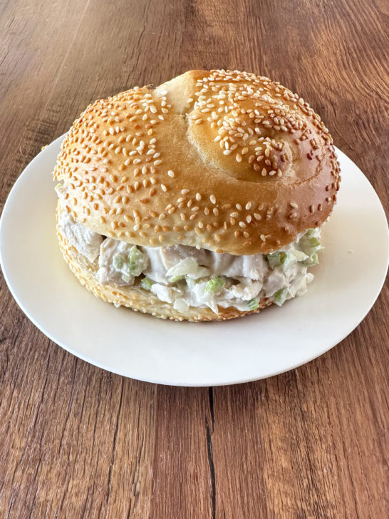 deli style chicken salad in a sesame seed roll on a white plate on a dark wood table.