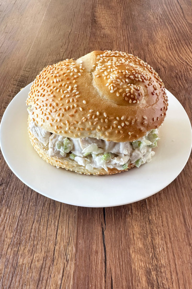 deli style chicken salad in a sesame seed roll on a white plate on a dark wood table.
