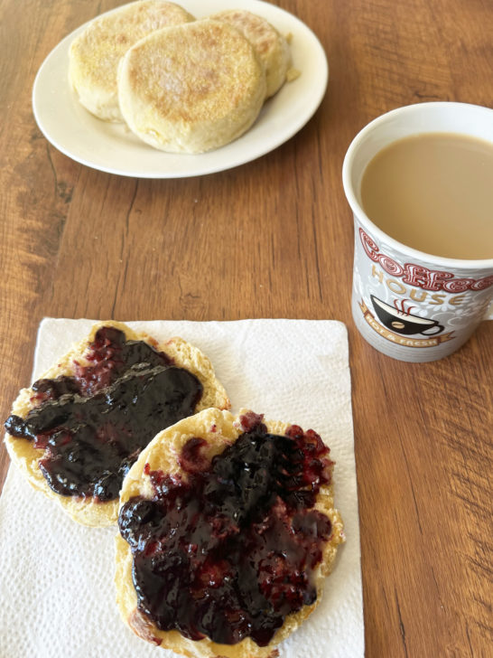 Dairy-free english muffins on a white plate in the background and a split english muffin with jelly on a napkin in the foreground next to a cup of coffee, all on a dark wood table