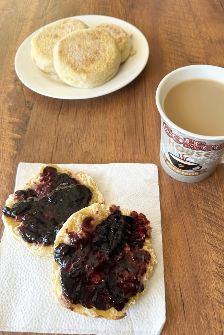 Dairy-free english muffins on a white plate in the background and a split english muffin with jelly on a napkin in the foreground next to a cup of coffee, all on a dark wood table
