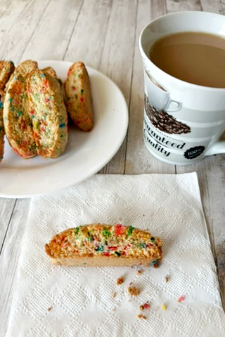 Funfetti cookie bars on a white plate with one funfetti bar on a napkin and a cup of coffee nearby on a white wood table.