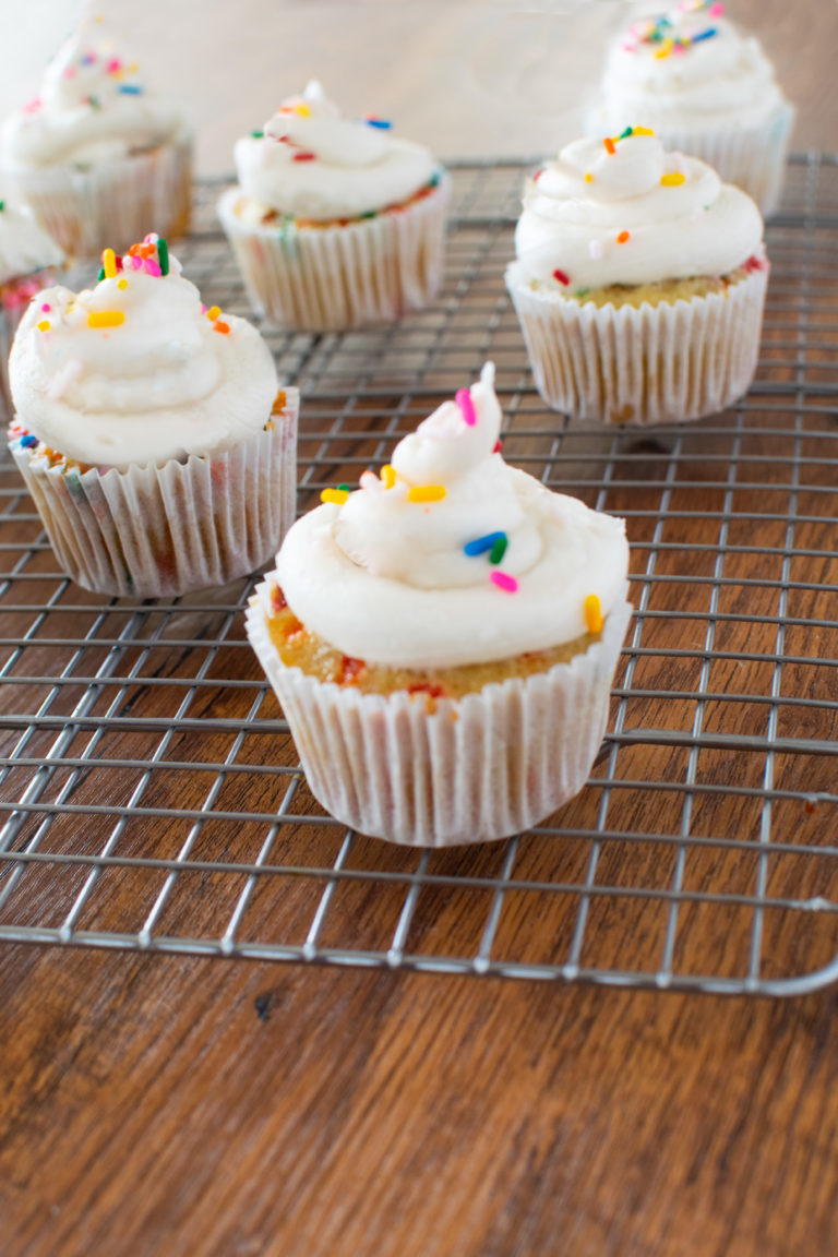 Homemade funfetti cupcakes on a cooling rack on a dark wood table.