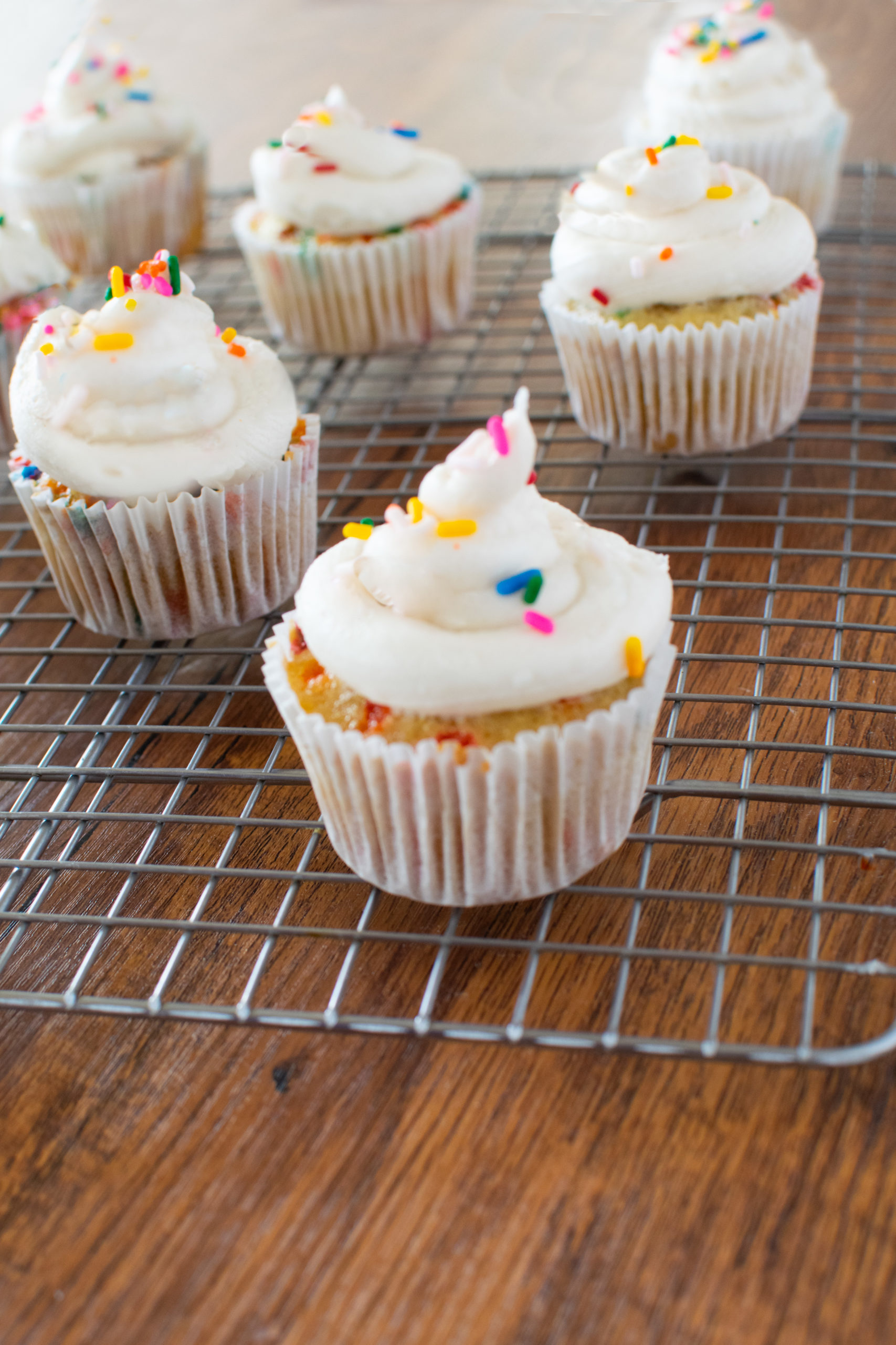 Homemade funfetti cupcakes on a cooling rack on a dark wood table.