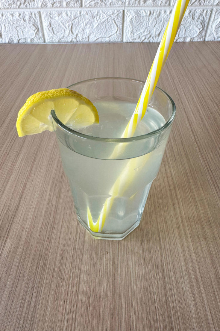 Clear glass of sweet lemonade with a slice of lemon and a yellow straw on a dark wood table