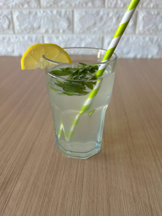 Limonana in a clear glass with a green straw and a slice of lemon and mint leaves floating at the top, on a wood table