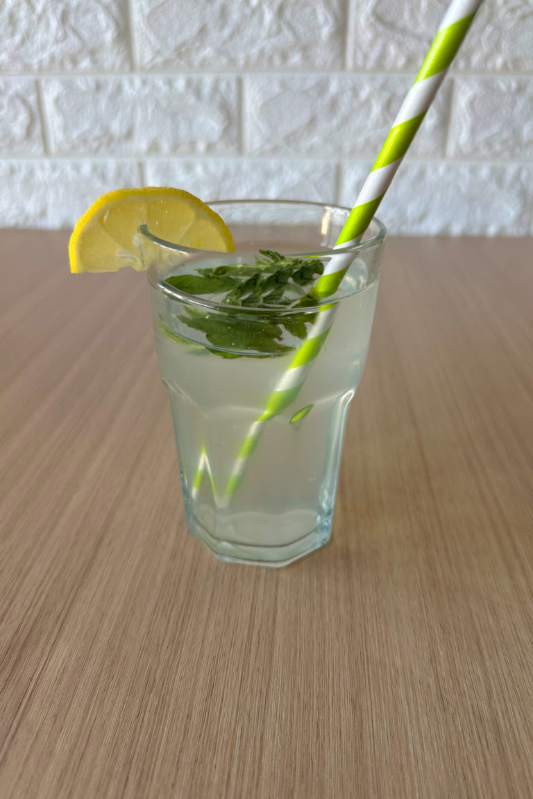 Limonana in a clear glass with a green straw and a slice of lemon and mint leaves floating at the top, on a wood table
