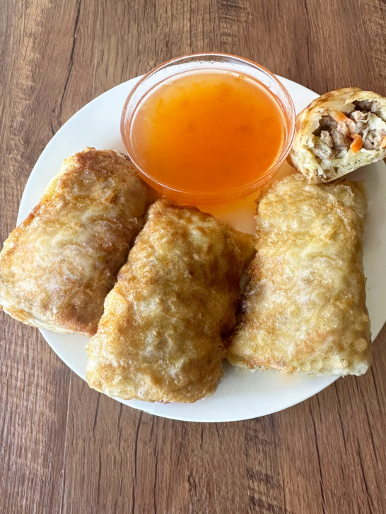 3 whole Beef egg rolls and one half egg roll on a white plate with a little clear bowl of duck sause behind them, all on a dark wood table