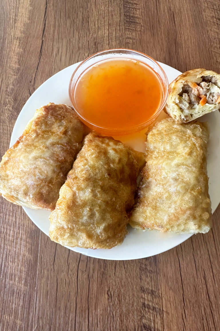 3 whole Beef egg rolls and one half egg roll on a white plate with a little clear bowl of duck sause behind them, all on a dark wood table