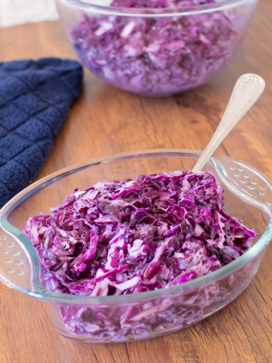 Red Cabbage Salad in a glass serving dish on a dark wood table with a blue napkin and a large bowl of the salad nearby