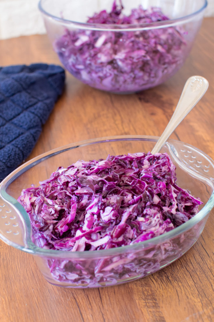Red Cabbage Salad in a glass serving dish on a dark wood table with a blue napkin and a large bowl of the salad nearby