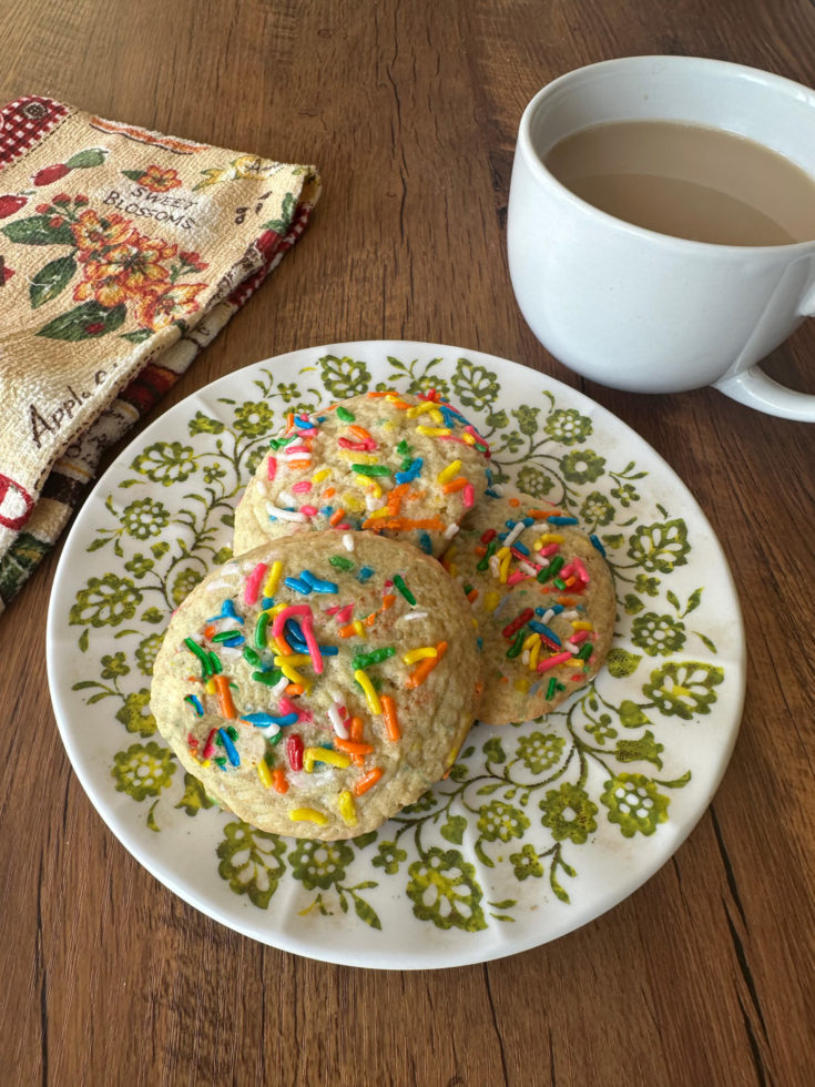 funfetti cookies on a green and white plate near a white mug of coffee and a towl on a dark wood table