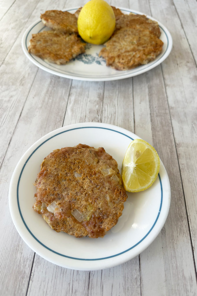 Tuna patty on a white plate wtih a slice of lemon and another plate of patties with a whole lemon in the background all on a white wood table