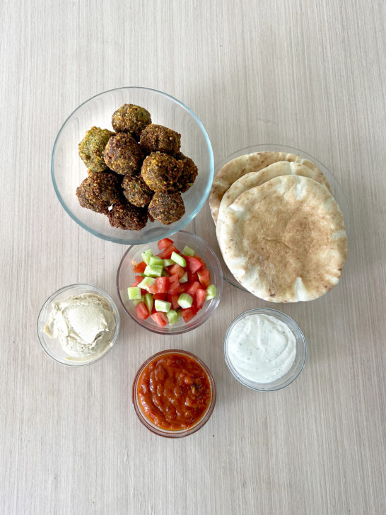 Felafel balls, israeli salad, hummus, tehini, matbucha all in clear bowls with a plate of pita nearby, all on a light wood table.