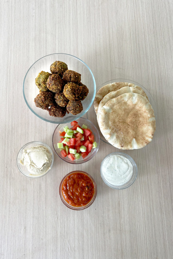 Felafel balls, israeli salad, hummus, tehini, matbucha all in clear bowls with a plate of pita nearby, all on a light wood table.