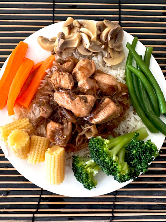 chicken chop suey on rice on a white place with sliced mushrooms, sliced carrots, long green beans, baby corn, and broccoli on a wooden table mat