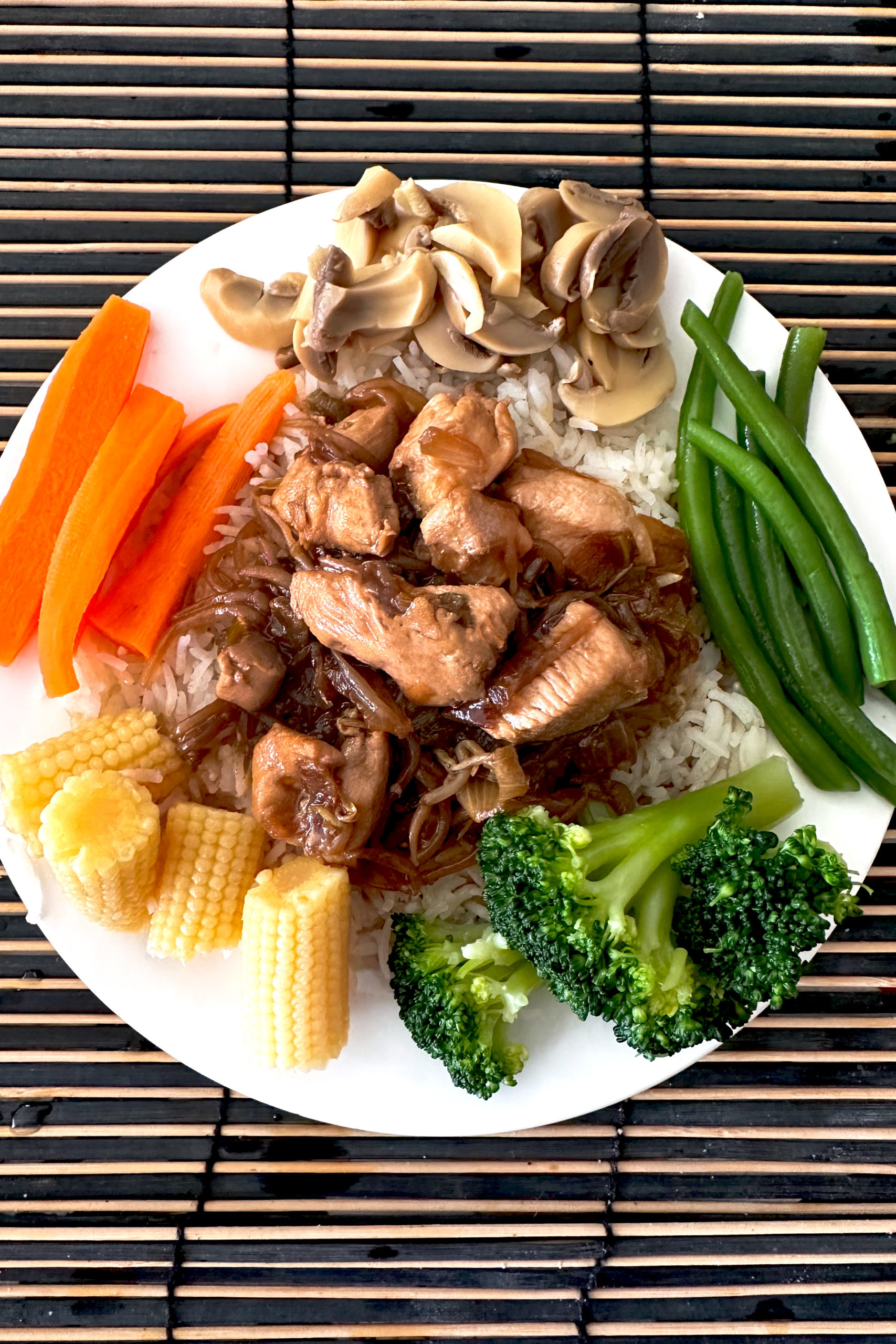 chicken chop suey on rice on a white place with sliced mushrooms, sliced carrots, long green beans, baby corn, and broccoli on a wooden table mat